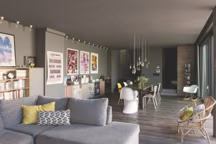 La pièce commune occupe presque tout le rez-de-chaussée. Au premier plan, un canapé chiné à L'Entrepôt, Marseille. Le décroché du plafond a permis de dissimuler les fixations des rideaux, d'intégrer les spots et les sorties de climatisation. Plancher en chêne, La Compagnie tropicale. Les piliers soutiennent en partie l'étage de la maison, dont la façade est montée en porte-à-faux. Les calligraphies qui les décorent ont été réalisées par l'artiste Tarek Benaoun, découvert au Mama Shelter de Paris. Rideaux en flanelle et velours réalisés sur mesure par Pascal Camberoque (atelierdudecorateur .com). Peintures Ressources. Dans le fond, mur monté en pierres sèches, Point P. Sur le mur de gauche, affiches de films érotiques des années 1970 trouvées chez le brocanteur spécialisé Homenibus, et affiche Dream Concert signée par l'artiste André, édition limitée, Kitsuné. Bibliothèque faite sur mesure par le menuisier Vladimir Drouzik à Marseille, console Ikea habillée de tissus choisis chez Eurodif, banquette en rotin chinée sur leboncoin.fr.