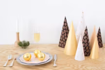 On aime la modernité de ce DIY centre de table de Noël imaginé par les talentueux Helen et Julien du blog Heju. Simples de formes mais décoratifs ces cônes rappellent l'esprit de Noël et de ses sapins mais dans une version graphique et contemporaine.

Pour ce DIY on a besoin de :

Des morceaux de tissus, un compas et de la colle, un rouleau de papier adhésif blanc utilisé habituellement pour faire des abat-jours.

Pour réaliser ces cônes en tissu et papier on commence par tracer un quart de cercle au verso du papier adhésif que l'on découpe ensuite. Avant de continuer on aura pensé à repasser les tissus à l'envers. En seconde étape on colle le quart de cercle découpé, coté autocollant, sur le tissu que l'on a choisi et on découpe l'ensemble. Puis avec la forme obtenue, désormais rigidifiée, on forme un cône dont on colle les bords.

Pensez à tracer plusieurs quarts de cercles de tailles différentes, à appliquer sur des tissus différents, pour obtenir des cônes de hauteurs et styles variés. A vous de jouer et de les rassembler, une fois terminés au centre de la table comme une forêt moderne de sapins géométriques.
Heju