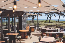 Bardage en tuiles de châtaignier teinté de RahuelBois, terrasse voilée de lin, tables en contreplaqué okoumé, chaises du Cartel de Belleville.